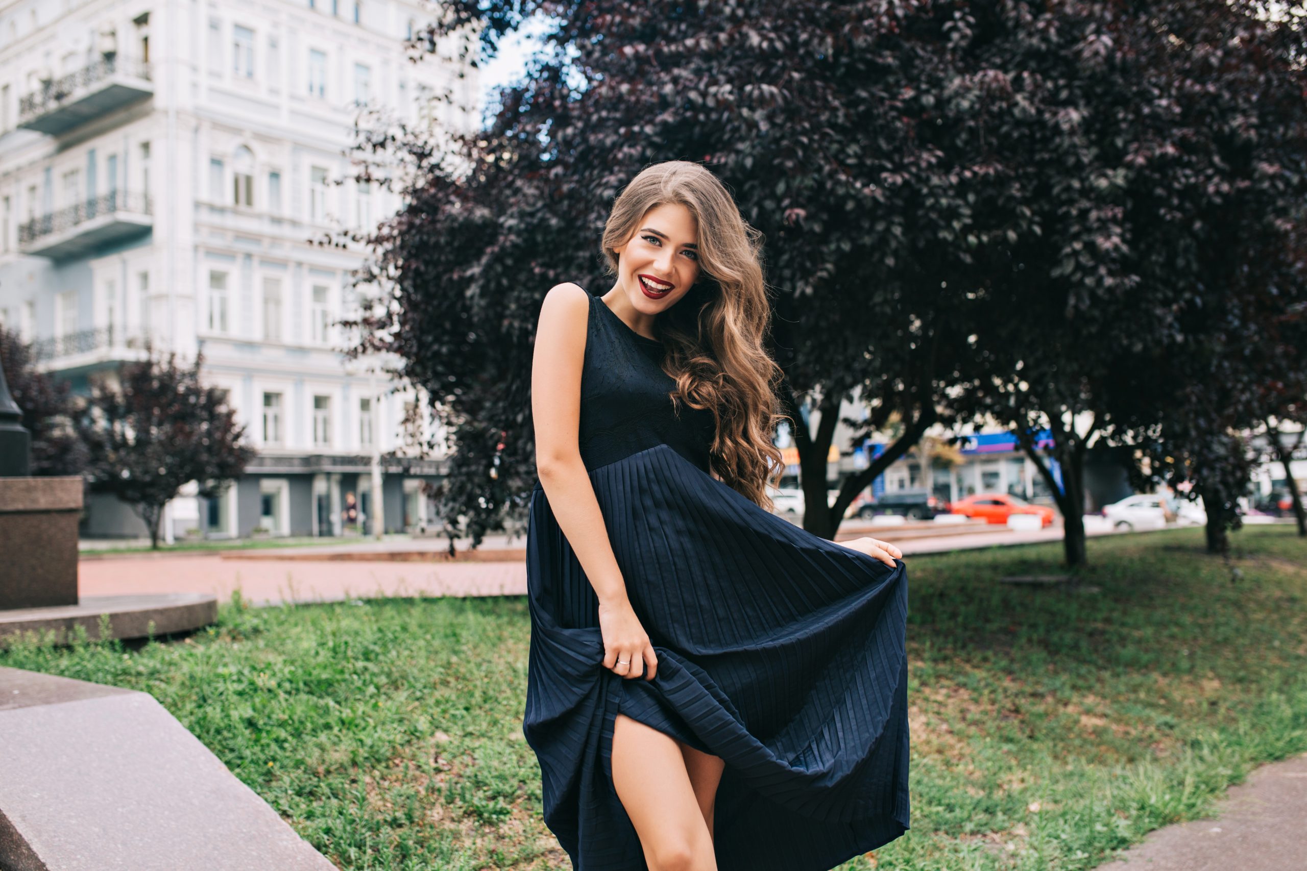 Elegant girl with long hair and vinous lips is posing in ciyu park. She holds long black dress and smiling.