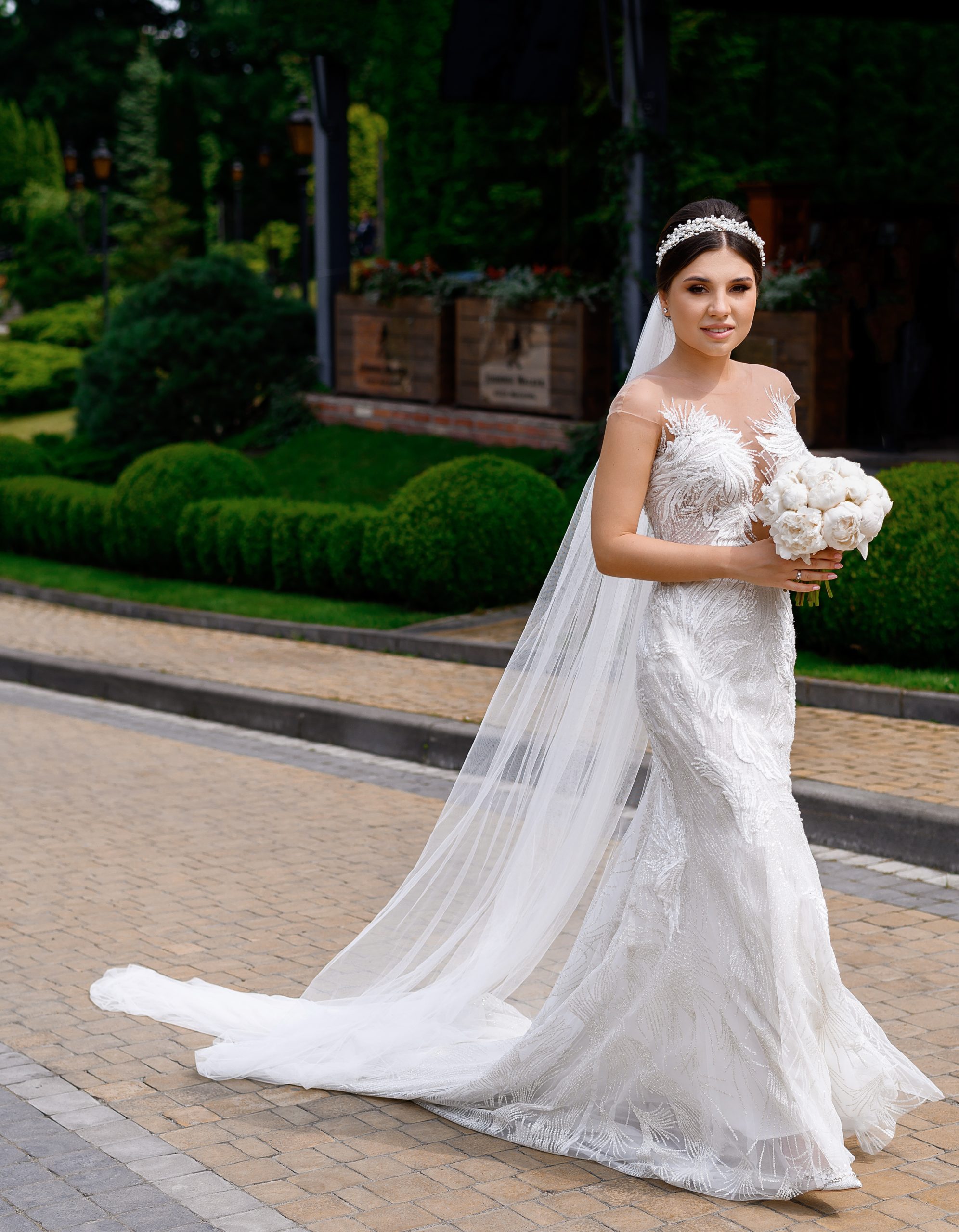 Front view of adorable woman with stylish hairdo, dressed in long veil and mermaid wedding dress with long trail, holding white peonies flowers, smiling and posing at camera on landscape background